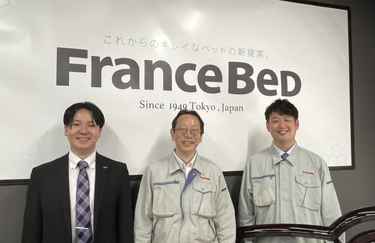 フランスベッド株式会社のロゴがある壁面に並んだ松尾英弥さんと伊橋孝明さんと山崎寛太郎さん
