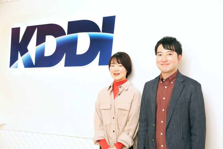 KDDI株式会社の前田さんと大橋さんが企業ロゴの前に並ぶようす