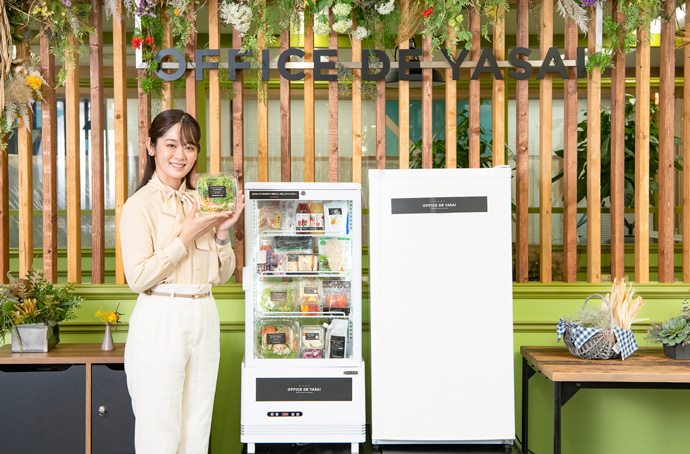 株式会社KOMPEITOの商品が納められる冷蔵庫の横で女性がサラダを持っている様子