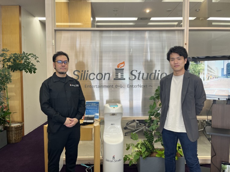 シリコンスタジオ株式会社の向井さんと江森さんが会社ロゴの前に並んで立つようす