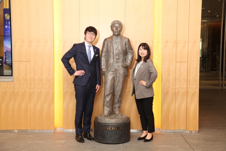 渋沢栄一像を挟んでポーズをとる商工会議所の逸見さんと松浦さん