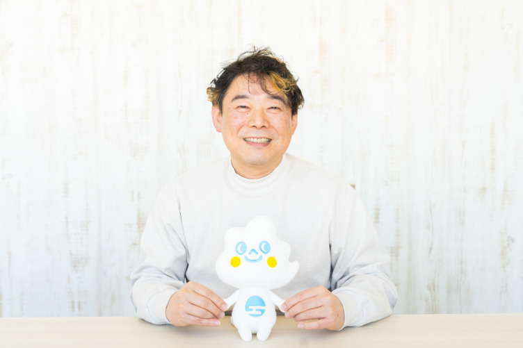トヨクモ株式会社の代表取締役社長である山本裕次さんが企業キャラクターのトヨクモちゃん人形を持って笑っている様子